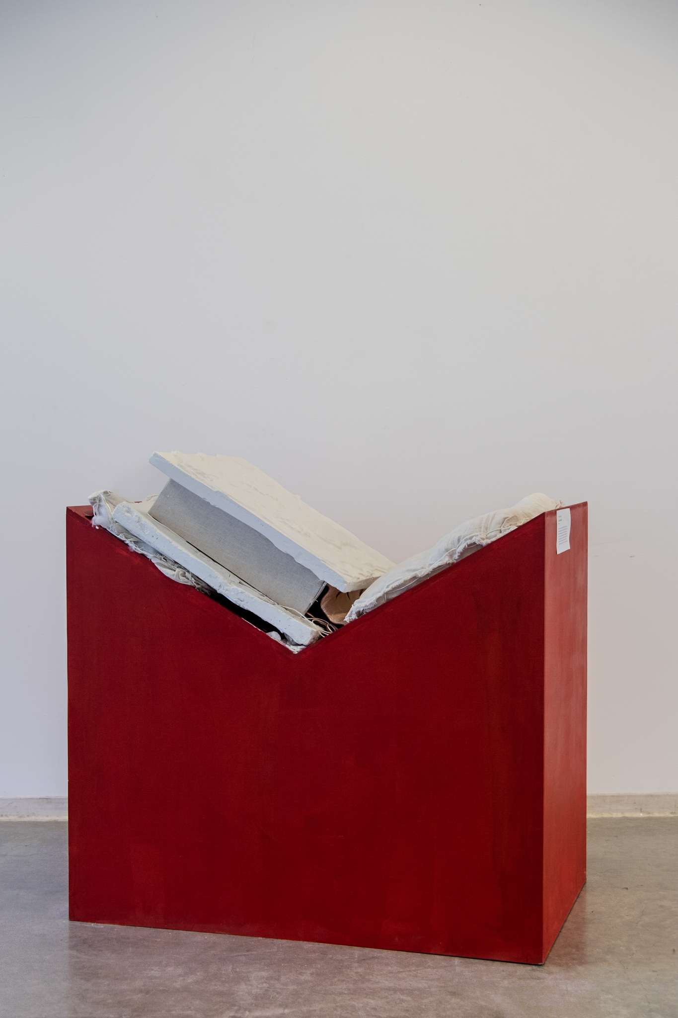 A red geometric sculpture with white and gray slabs forming a book protruding from the top, displayed against a plain white wall.